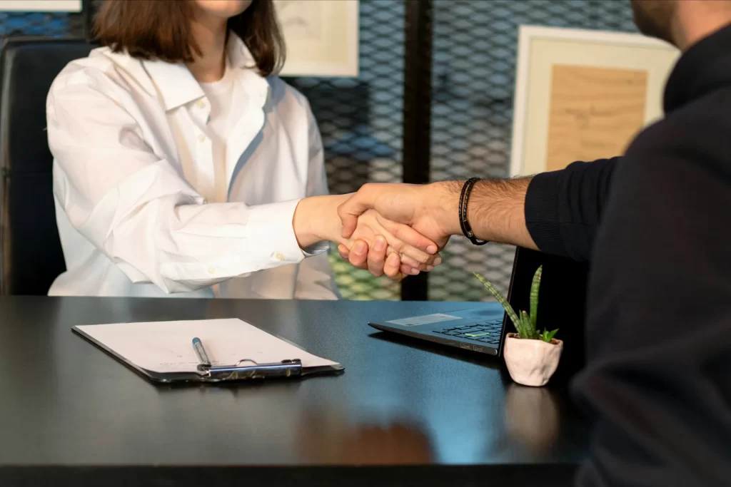 Two people shaking hands across a desk during a professional job interview, symbolizing a successful hiring process. A notepad and resume are visible on the table. Recruitment agency Scotland supports connections like this.
